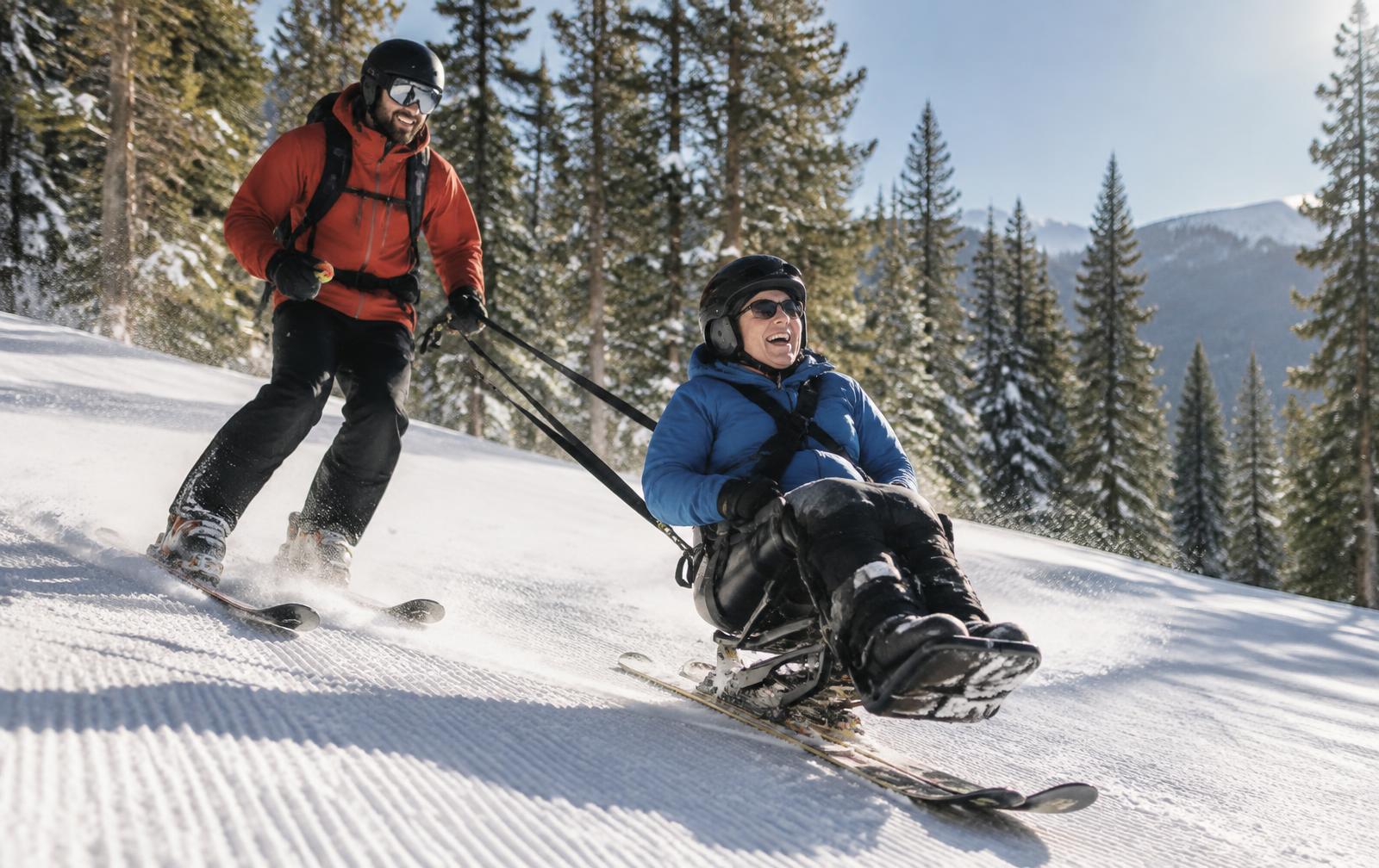 An adaptive skier in a sit-ski laughing as an instructor skis alongside on a sunny forest trail
